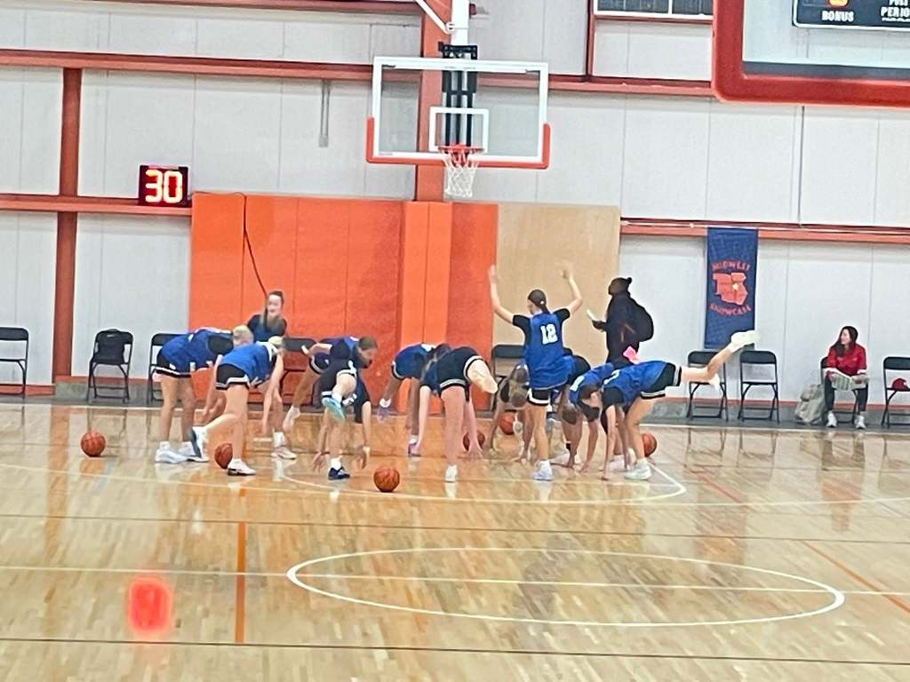 A basketball team in blue uniforms practicing on the court, bending down to pick up basketballs. A scoreboard shows a 30-second timer in the background.