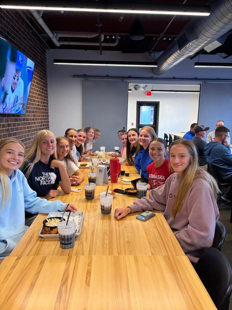 A group of young women sitting together at a long wooden table in a casual dining setting, enjoying meals and drinks, with a television mounted on the wall displaying a sports game in the background.