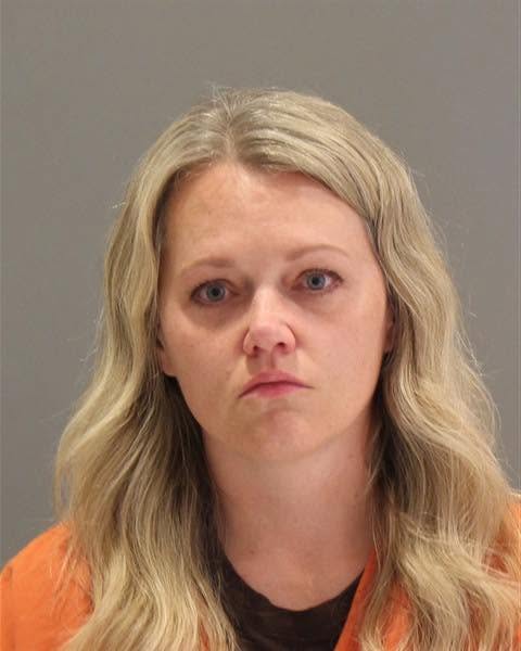 A woman with long, wavy blonde hair wearing an orange jail uniform, looking directly at the camera with a neutral expression.