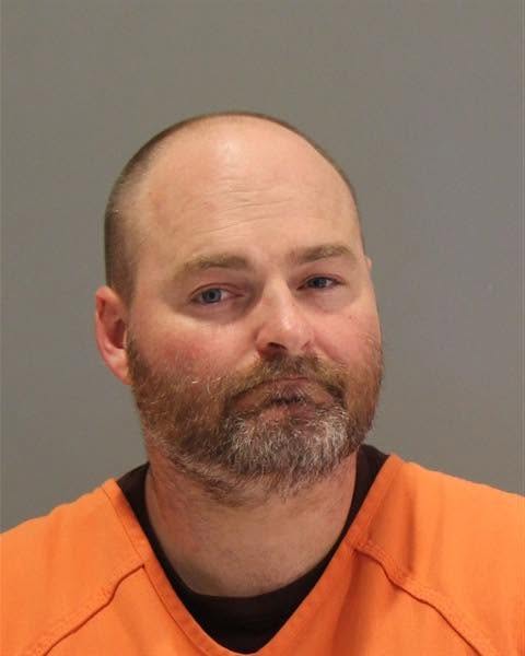 Mugshot of a man with a shaved head and a beard, wearing an orange prison jumpsuit, against a gray background.