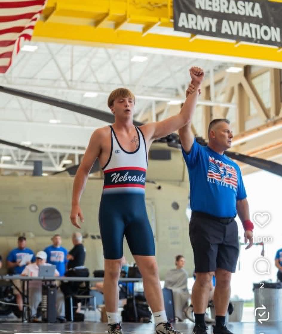 A young male wrestler in a Nebraska singlet celebrates his victory with his hand raised by a coach in a blue t-shirt, surrounded by spectators inside a large venue.
