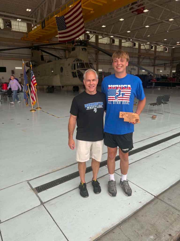 Two men standing together inside a large hangar with an American flag and a military helicopter in the background. One man is holding an award and wearing a blue shirt, while the other is wearing a black shirt.