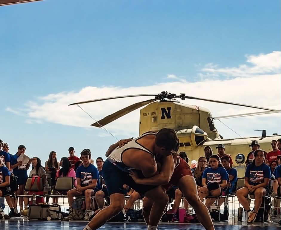 Two wrestlers engage in a match on a wrestling mat, with a military helicopter visible in the background and a crowd watching.