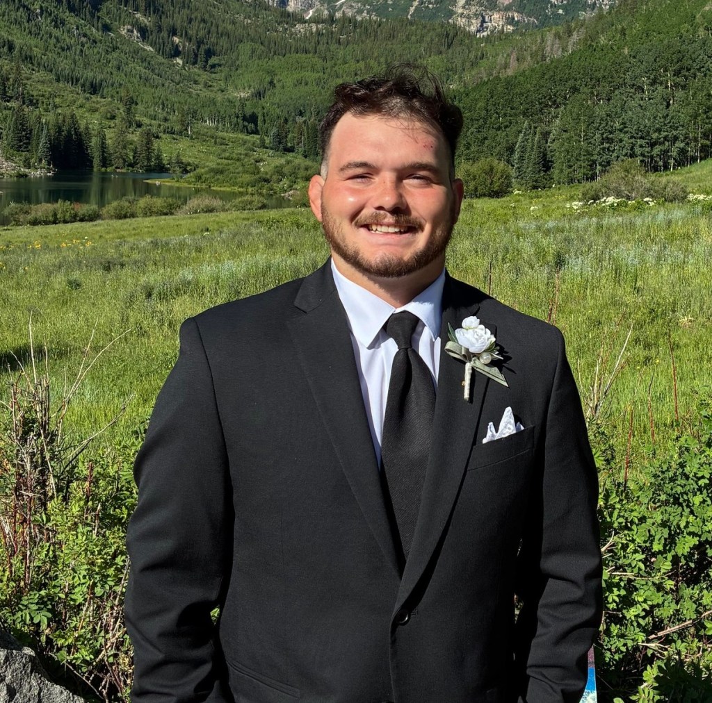 A man wearing a black suit and tie, smiling brightly, stands in a lush green outdoor setting with mountains in the background and a small lake visible.