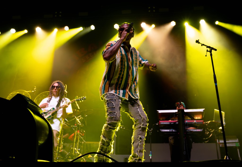 A male singer performing live on stage with a microphone, wearing a colorful striped shirt and ripped jeans, in front of a vibrant yellow light backdrop. A guitarist is visible in the background, playing a guitar.