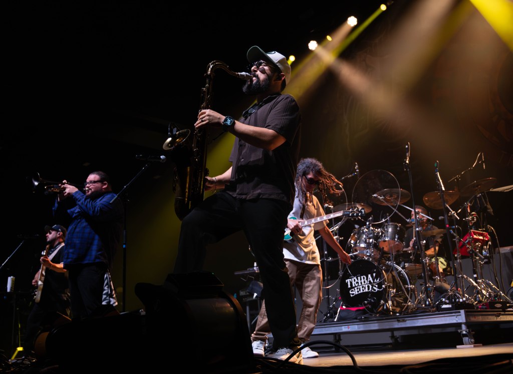 A live music performance featuring a band with a saxophonist, trumpeter, and other musicians on stage, illuminated by stage lights.