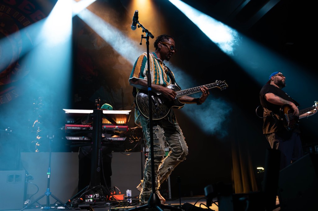 A musician playing an electric guitar on stage with bright lights and a band member on keyboard in the background.