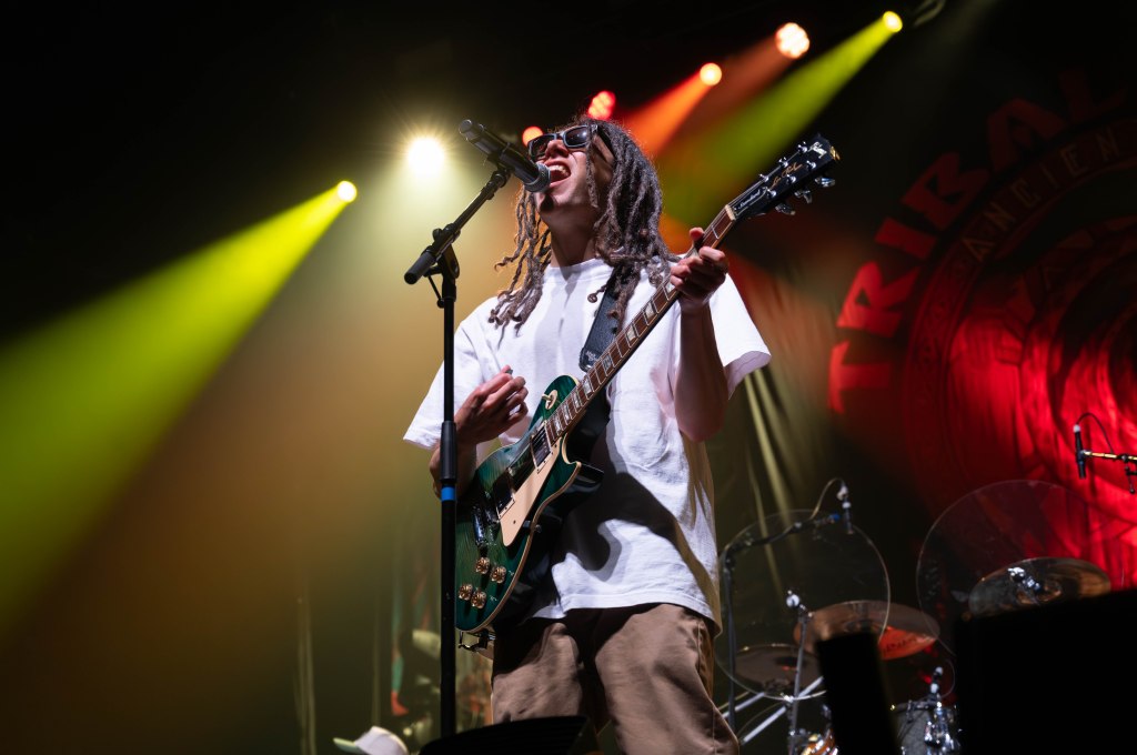 Musician performing on stage with a guitar, wearing sunglasses and a casual outfit, under colorful lights.