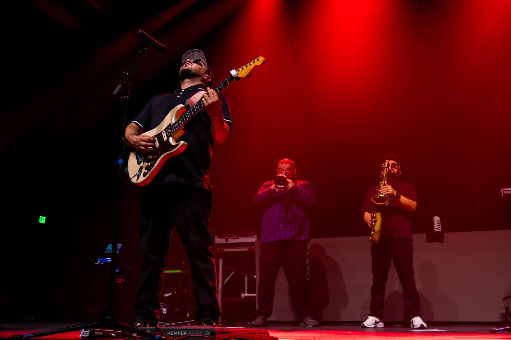 A musician playing an electric guitar on stage with two brass players in the background, illuminated by red stage lighting.