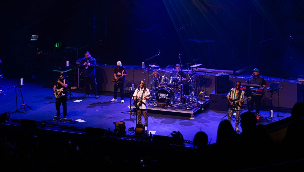 A live band performing on stage, featuring multiple musicians with instruments, including a guitar and drums, under stage lights.