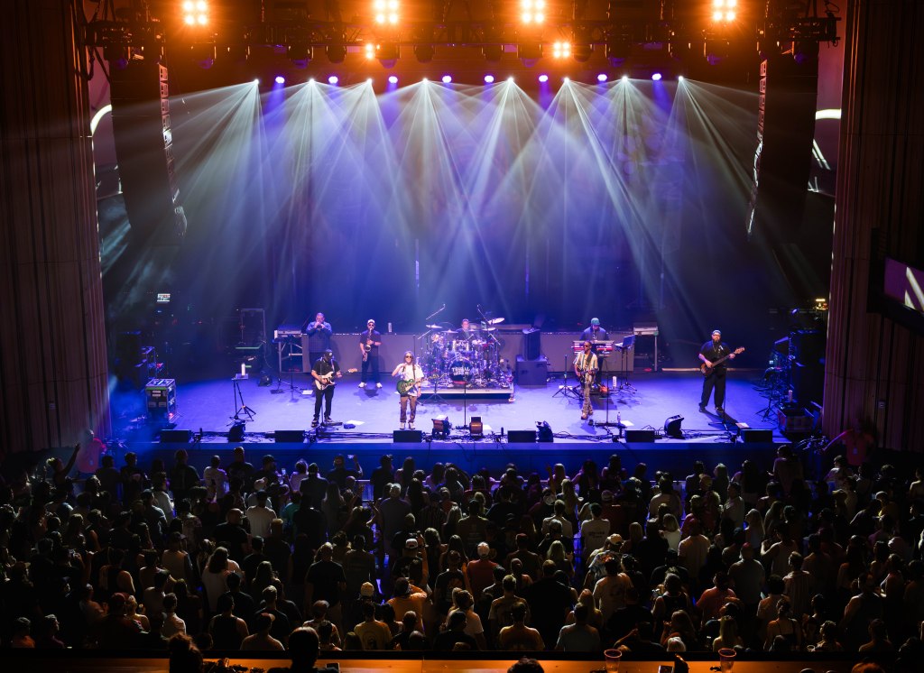 A live concert scene with a band performing on stage, illuminated by colorful lights, and an audience watching from below.