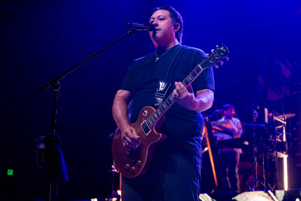 A musician performing on stage with an electric guitar, microphone in hand, under colorful stage lights.