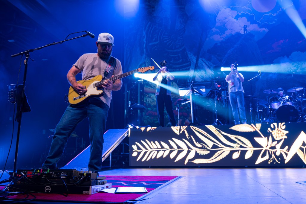 Guitarist performing on stage with a band, featuring brass musicians and a drummer, under vibrant stage lighting.