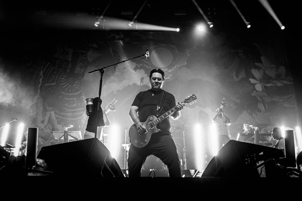 A guitarist performing on stage in a black and white setting, surrounded by musicians and stage lights.