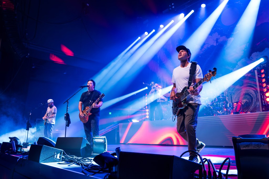 A band performing on stage with bright stage lights and smoke effects. The musicians include a guitarist, a bassist, and a singer, creating an energetic atmosphere.