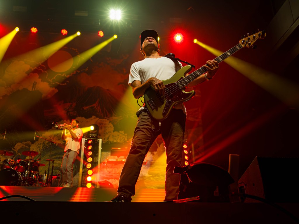 A musician playing a bass guitar on stage with dramatic lighting in a concert setting, accompanied by another musician in the background playing a trumpet.