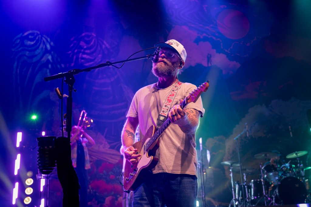 A guitarist performing on stage with colorful lighting and a band in the background.