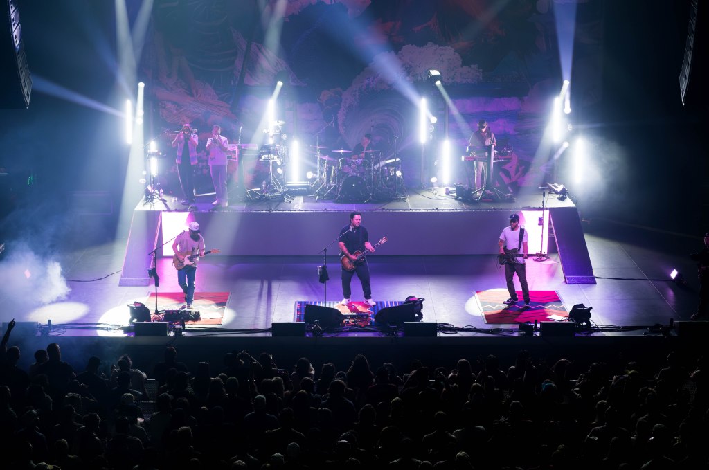 A live concert scene featuring a band performing on stage with dramatic lighting and a crowd of fans in front.