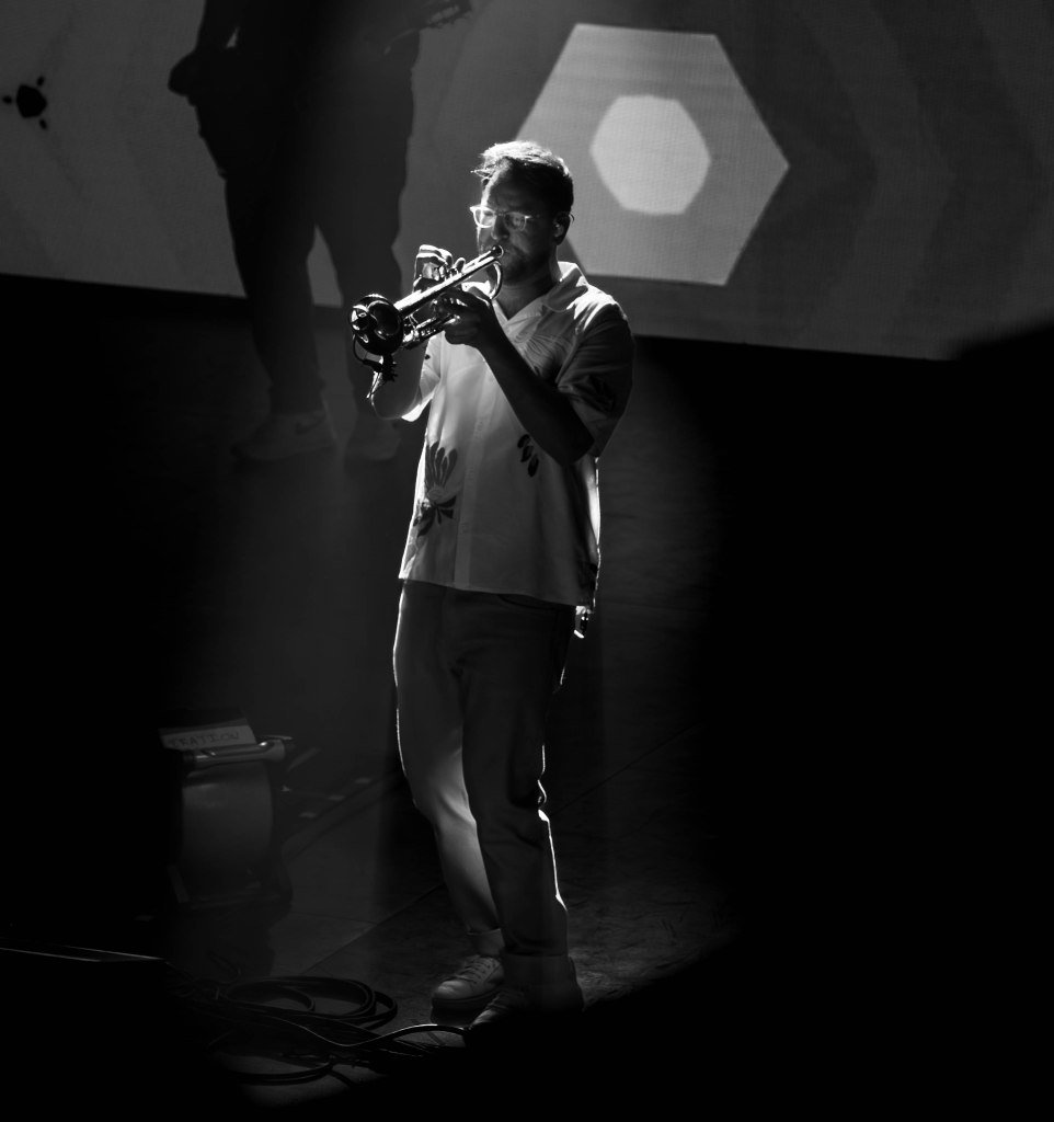 A musician playing the trumpet on stage, illuminated by spotlight, in a black and white setting.