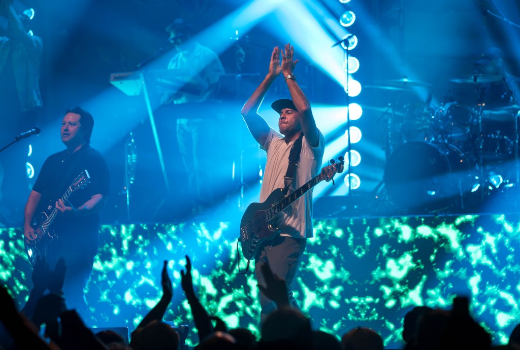 A live band performing on stage, featuring a guitarist and a bass player with their hands raised. Colorful lights and a dynamic visual backdrop create an energetic atmosphere.