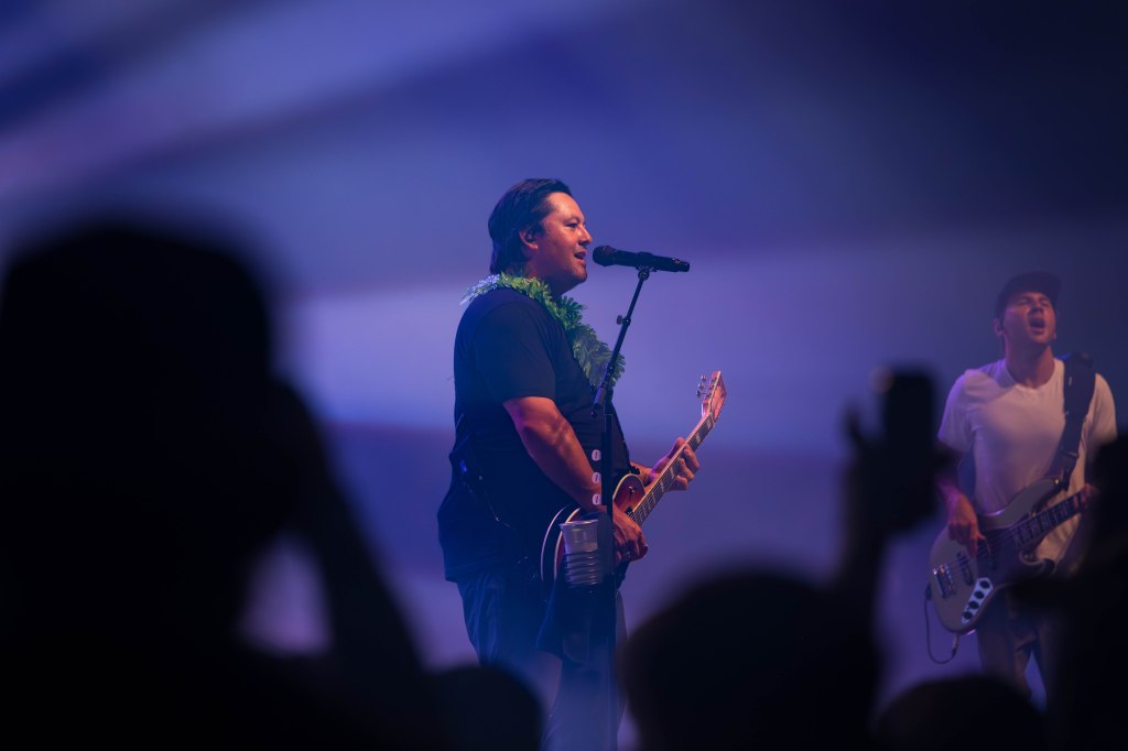 A musician performing on stage, singing into a microphone while playing an electric guitar, with a band member visible in the background playing bass. The stage is lit with colorful lights.