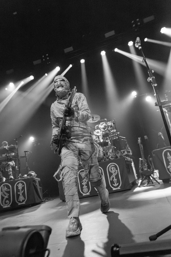 A musician dressed in a mummy costume performing on stage, holding a guitar, with bright stage lights illuminating the scene.