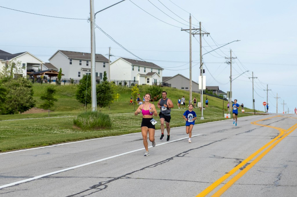 A group of runners participating in a race on a suburban road, with houses and green grass in the background.