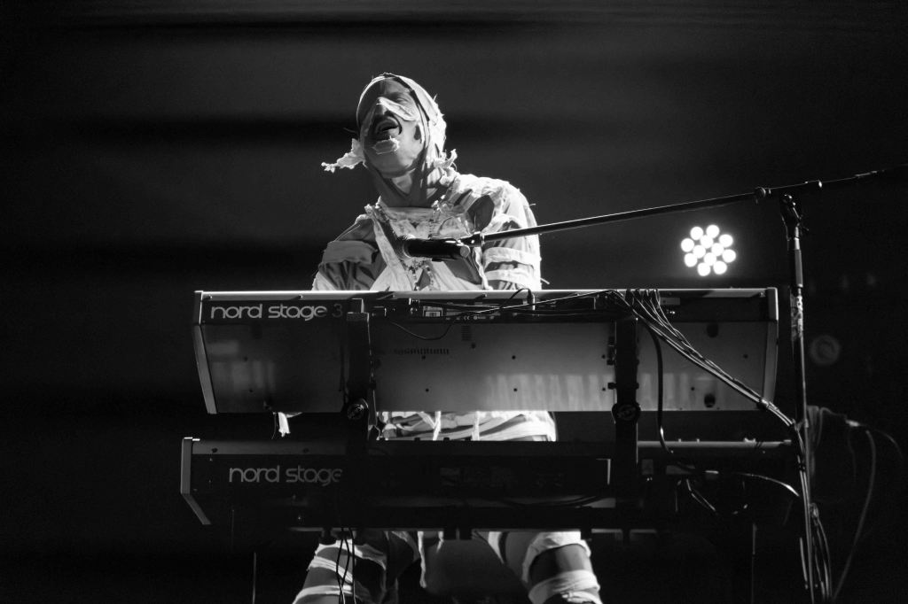 A musician performing on stage, wearing a white outfit and playing a keyboard, with dramatic lighting and a microphone.