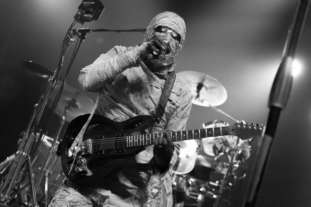 A musician dressed in elaborate, tattered clothing and a wrapped mask plays an electric guitar on stage, with a drummer in the background.