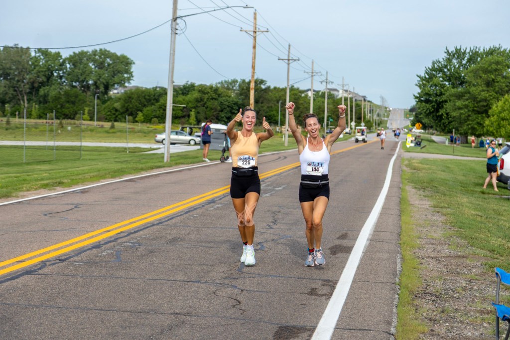 Two female runners celebrating as they approach the finish line of a road race, with a rural backdrop and spectators in the background.