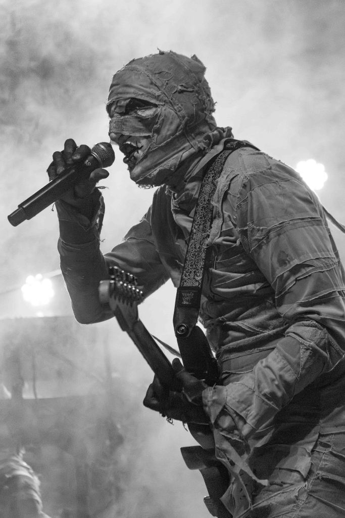 A musician dressed in a mummy costume performing on stage, holding a microphone and electric guitar, surrounded by smoke and dramatic lighting.