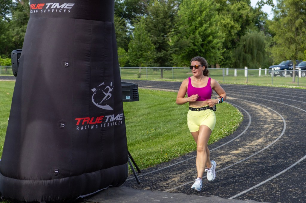 A woman is jogging on a track wearing a pink tank top and yellow shorts, with sunglasses and a fitness watch. In the background, there is a large inflatable arch with 'TRUE TIME RACING SERVICES' branding.