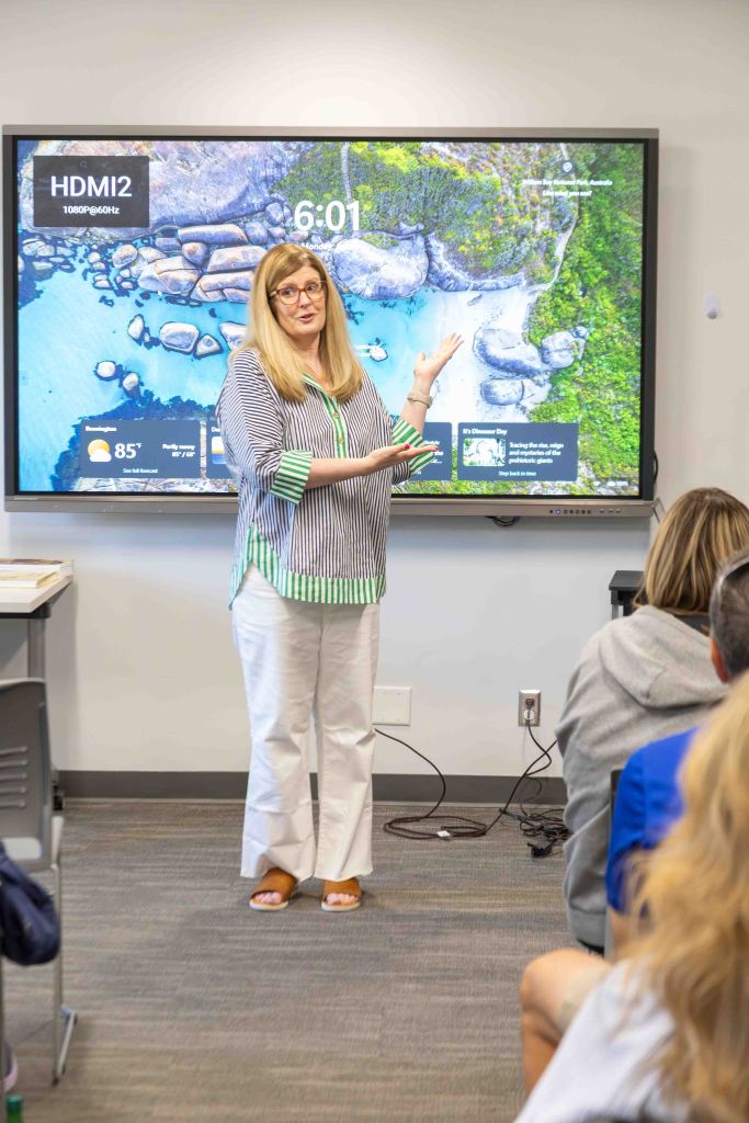 A female presenter stands in front of a large screen displaying a scenic beach view, discussing a topic while pointing at the screen. Audience members are seated in the background.