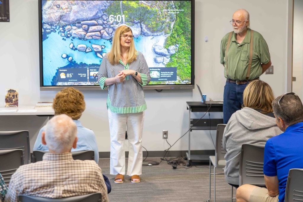 A woman speaking to an audience in a room with a large screen displaying a scenic image and weather information. An older man stands beside her, while attendees listen attentively.
