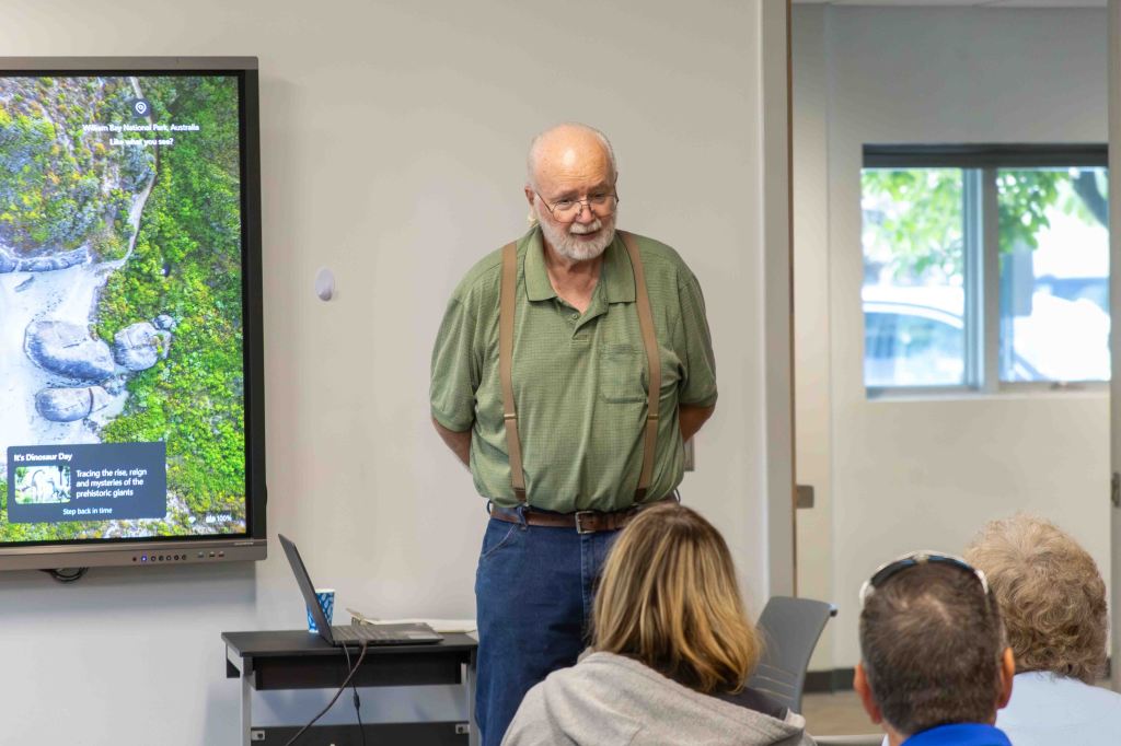 An elderly man with glasses and suspenders stands in front of a digital screen displaying a map of a national park, while a small audience listens attentively.