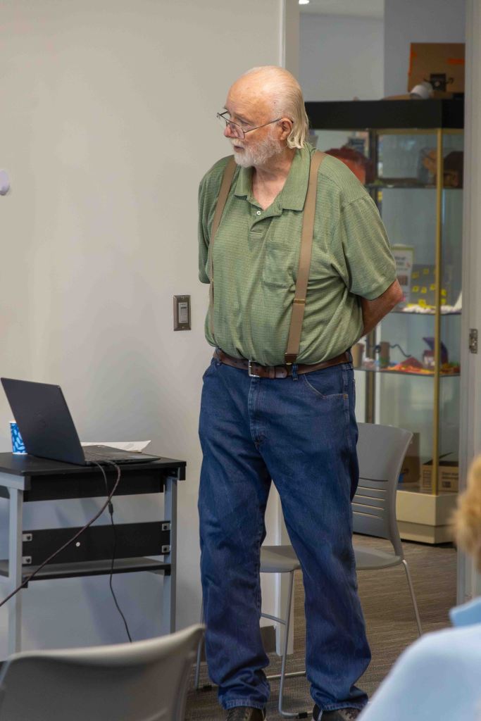 An elderly man with glasses and white hair stands next to a laptop on a table, wearing a green shirt and blue jeans.
