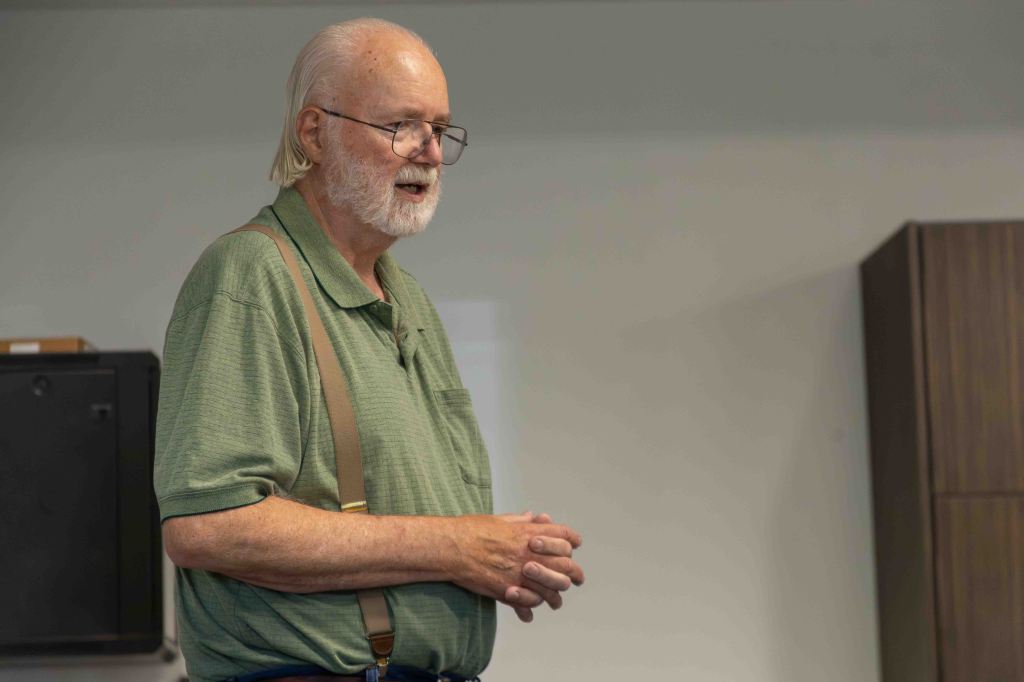 An older man with glasses and long white hair stands speaking in front of a classroom setting, wearing a green shirt and suspenders.