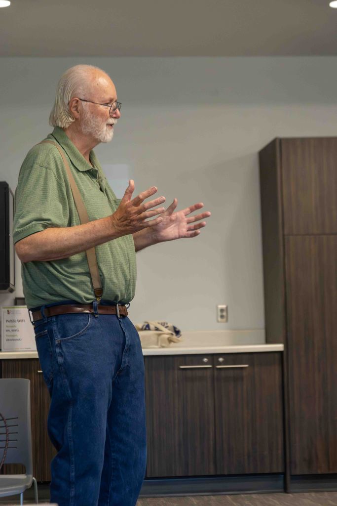 An older man with a beard and glasses is standing and gesturing while speaking, wearing a green shirt and suspenders, in a modern kitchen or meeting space.