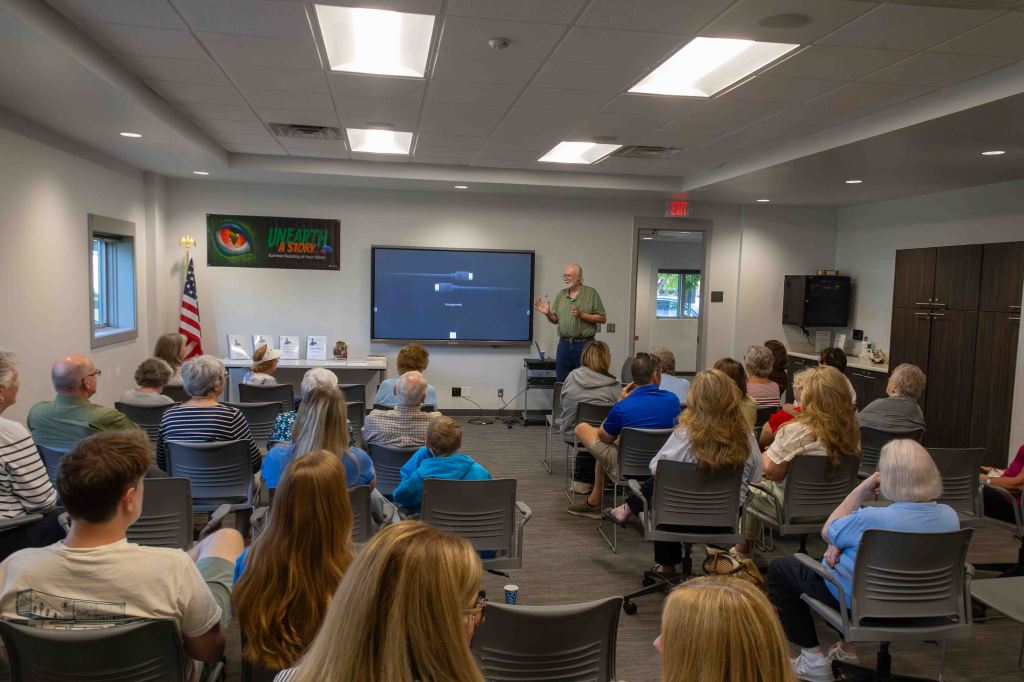 A speaker presents to an audience in a modern indoor space, featuring a large screen displaying a presentation, with attendees seated in rows and various seating arrangements.