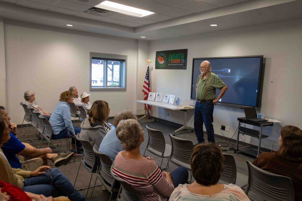A speaker presenting to an audience in a meeting room, with chairs arranged facing a display screen and a table displaying materials.