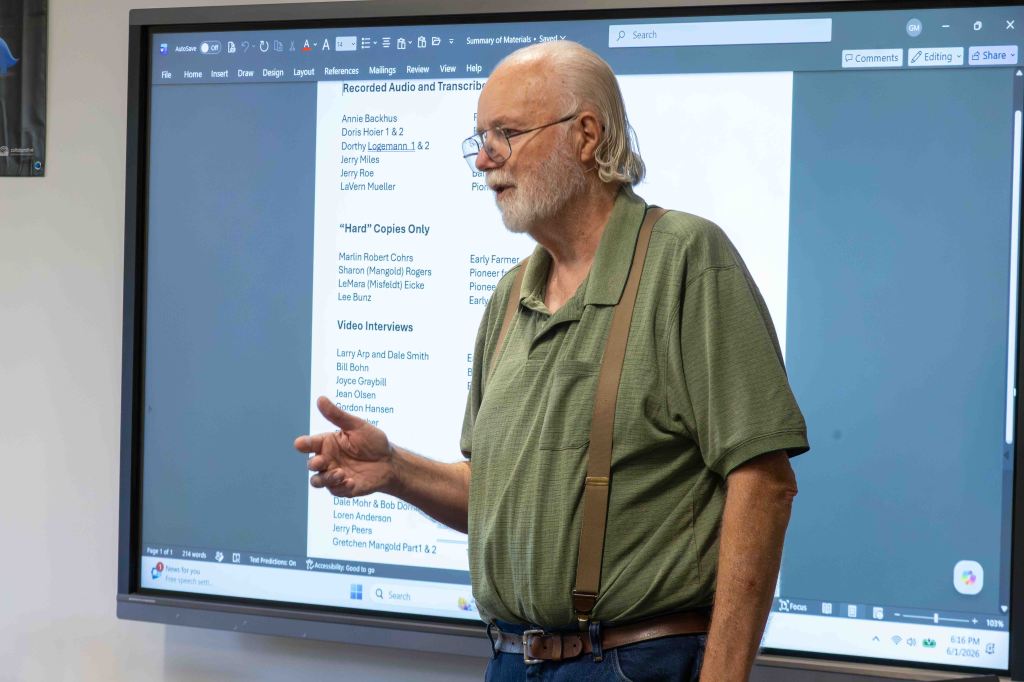 A man with glasses and long hair, wearing a green shirt and suspenders, stands in front of a digital screen displaying a list of names and topics related to recorded audio and video interviews.