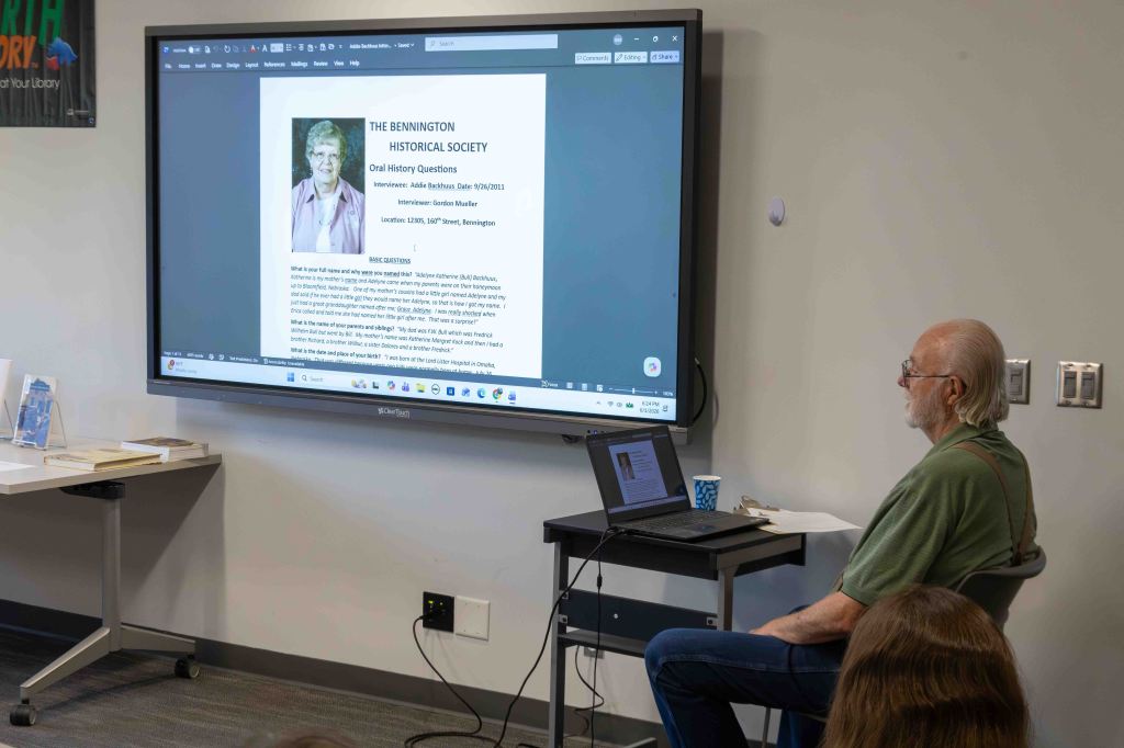 A person sitting at a table in front of a large screen displaying a document titled 'The Bennington Historical Society' with oral history questions. The individual is wearing glasses and has long hair, while a laptop and paper are placed on the table.