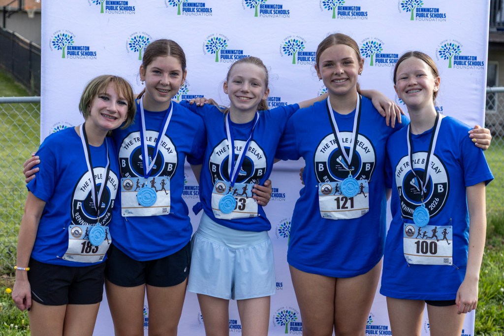 Five athletes in blue t-shirts pose together, smiling and wearing medals after a relay race, with a backdrop featuring the Bennington Public Schools Foundation logo.