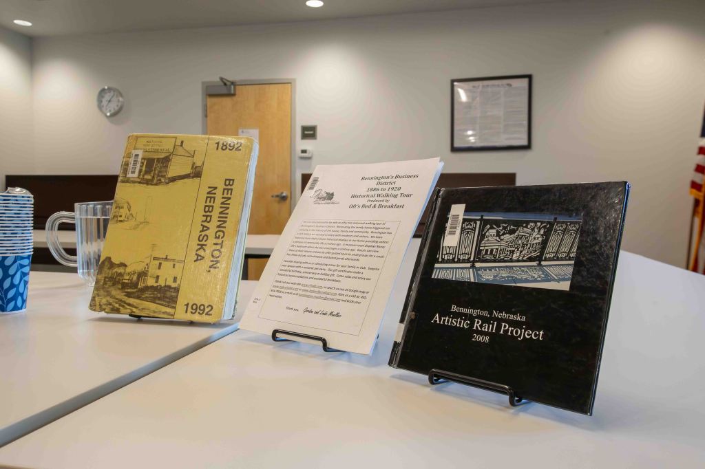 A table display featuring three items: a yellow booklet titled 'Bennington, Nebraska 1992,' a white paper about Bennington's business district walking tour, and a black booklet titled 'Bennington, Nebraska Artistic Rail Project 2008,' alongside a glass pitcher and a blue cup with a patterned design.