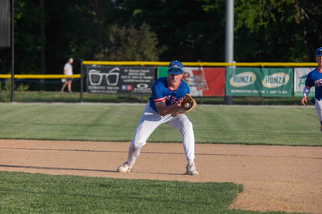 A young male baseball player in a blue and red uniform is crouched in a fielding position on a baseball diamond, preparing to catch a ball, with another player nearby.
