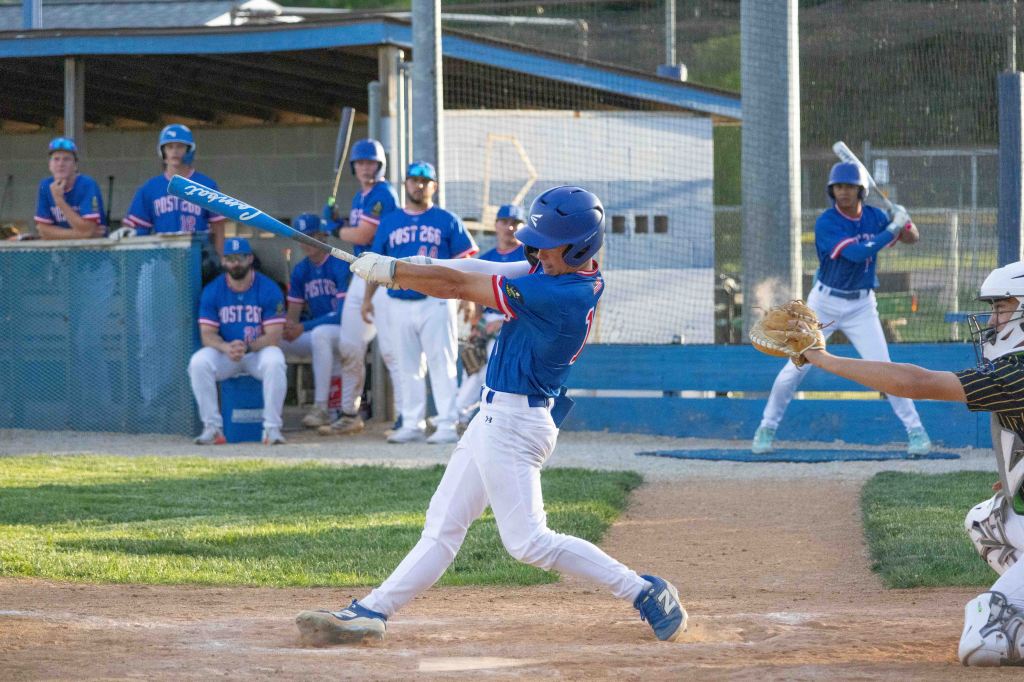 A young baseball player swings a bat during a game, while teammates watch from the dugout. The scene captures the moment of impact as the player makes contact with the ball.