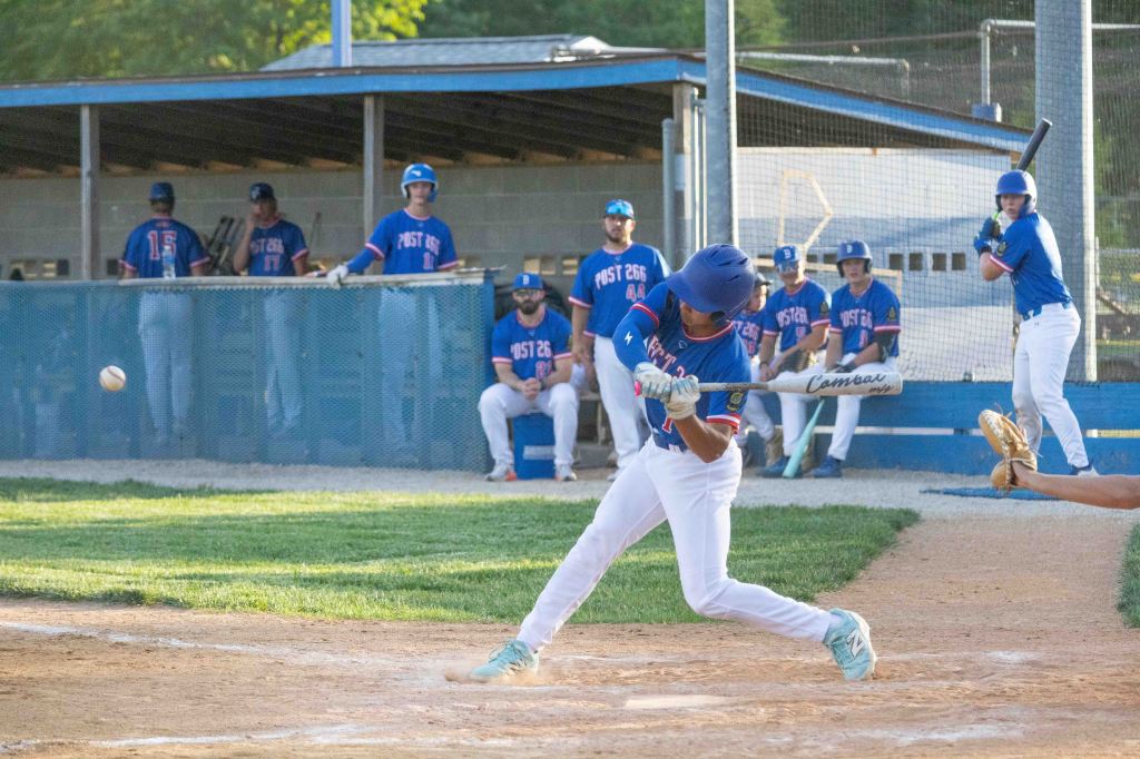 A young baseball player swings at a pitch while playing in a game, with teammates seated in the dugout in the background.