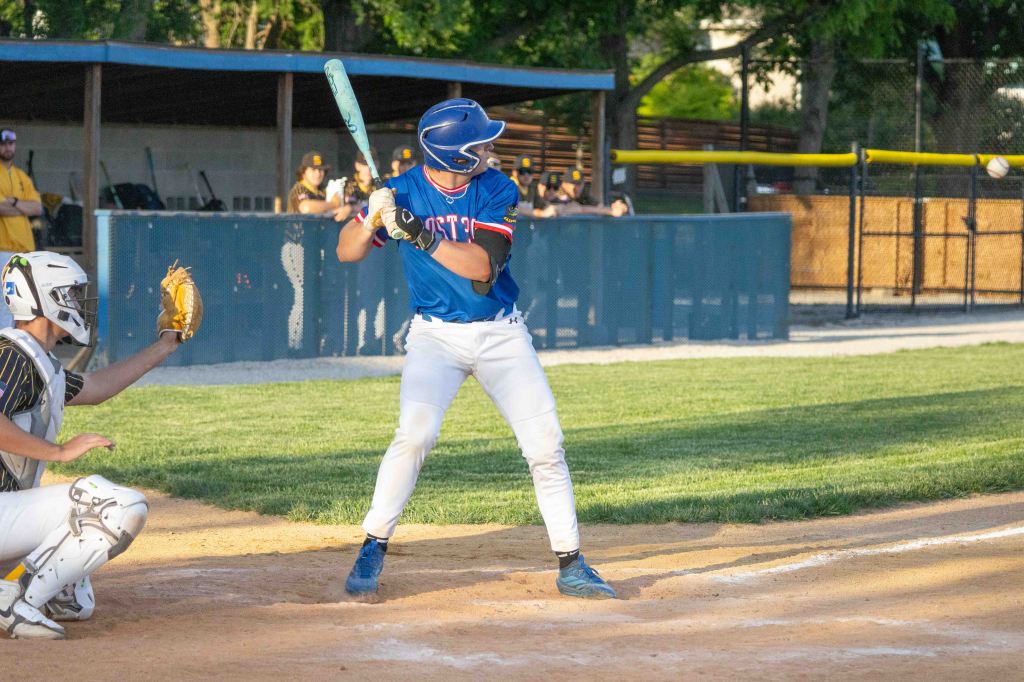 A baseball player in a blue jersey prepares to bat while a catcher in a white uniform crouches behind home plate during a game.