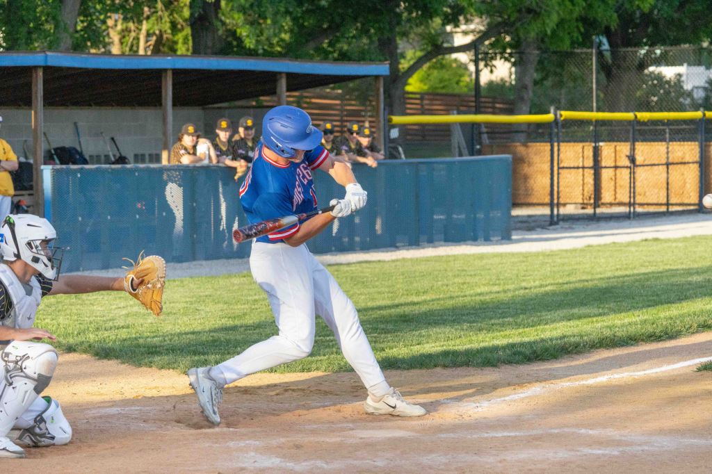 A baseball player in a blue and red uniform swings a bat at a pitch while a catcher crouches behind home plate.