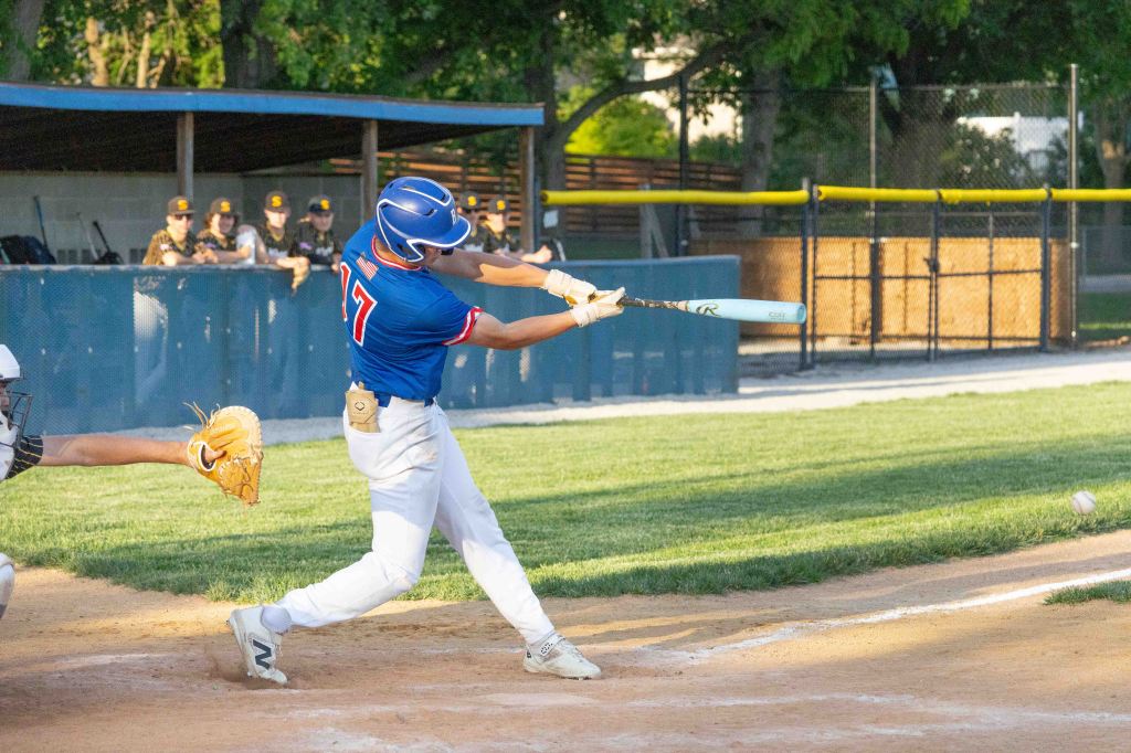 A baseball player in a blue jersey swings a bat at a pitch in a sunny outdoor field, with spectators in the background.
